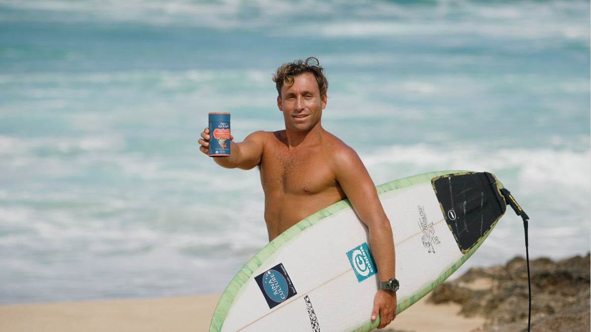 Surfer holding AiNA culture turmeric, cacao elixir blend on the beach in Hawaii with surfboard 